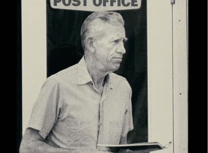 A black-and-white photo of J.D. Salinger's grim-looking side profile. He stands underneath a post office sign and holds a slim book.