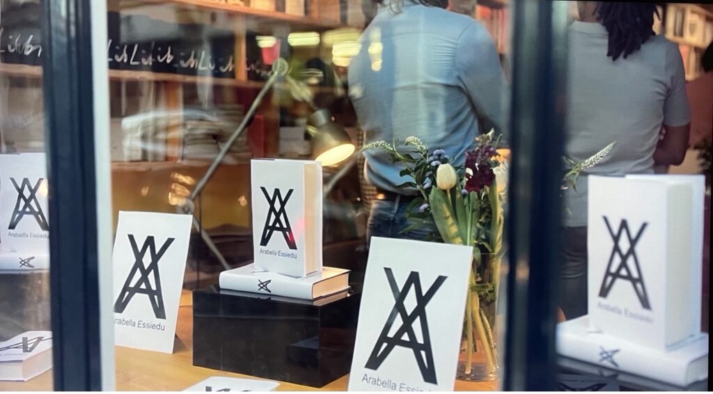 A bookstore window that showcases several copies of Arabella's book. The book covers are white with a large A with an X intersecting the letter and Arabella Essiedu's name written underneath. People are in the background milling around the store.