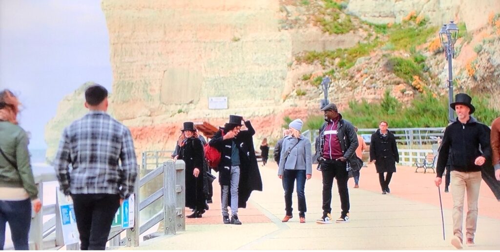A group of people congregate on a boardwalk, with cliffs in the background. Others walk past. Some wear top hats.