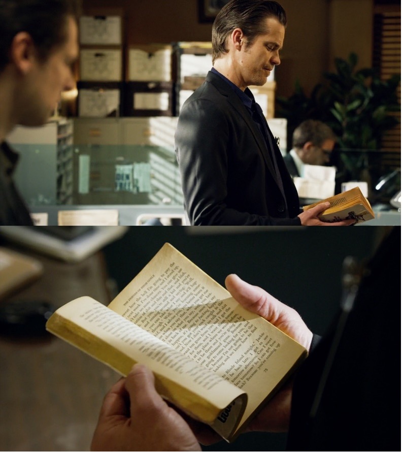 A man in an office is holding and examining a paperback book. Another man watches him.