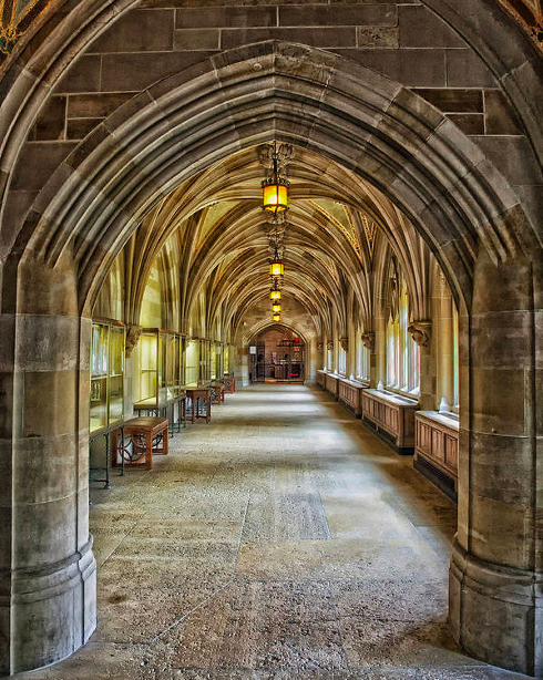 A hallway in a Gothic building lined by cement pillars under an arched roof. The left side of the hallways shows a series of small tables.