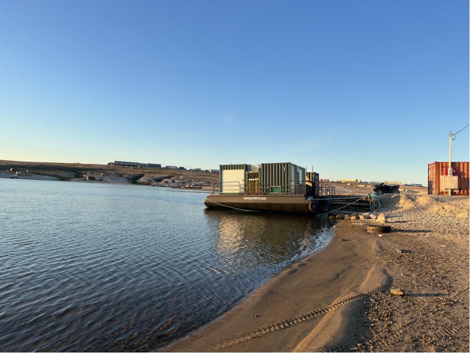 A photo of a bay shoreline, with a sandy beach crossed with ATV tracks in the foreground, and a research barge moored to the shore in the center of the image. The barge is labeled Qiniqtiryuaq and three green shipping containers are visible on its deck. Modular buildings are visible in the background on the ridges of Gjoa Haven.]