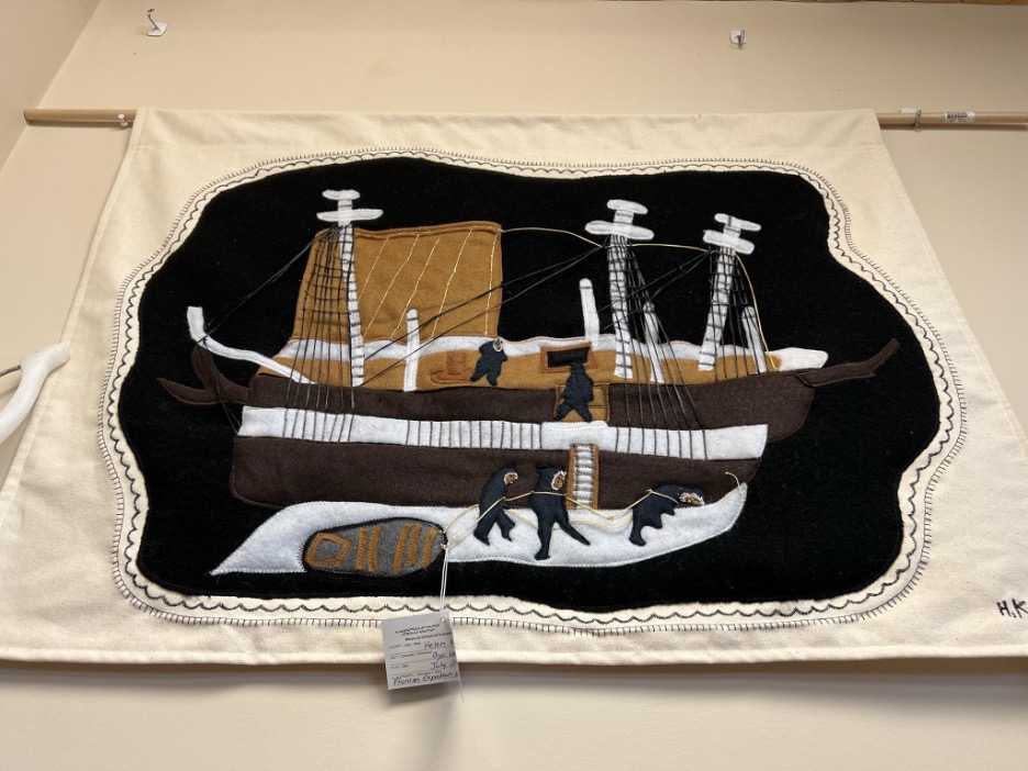 a photo of a large felted and embroidered wall tapestry hanging from a wooden rod. The background is cream-colored canvas, and on a field of black, with brown, white, and tan accents, the artist Helen Kaloon has represented the icebound wooden ship Erebus, its masts slightly askew and a tarp still raised over its deck for overwintering; the shrouds are rendered three dimensionally. A party of five Inuit are depicted taking usable items from the ship and carrying them away on a sled that had been loaded heavily enough that one Inuk is shown half-bent from the weight.]