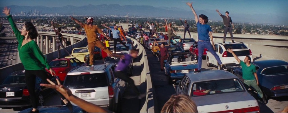 people in brightly colored outfits dance atop cars stopped in traffic on the freeway